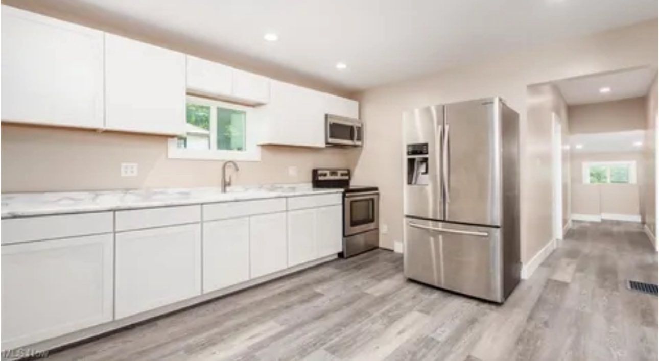 A bright kitchen with white cabinets, stainless steel appliances, marble-look countertops, and gray wood-plank flooring.