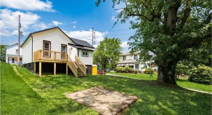 A white cottage with a raised wooden deck and stairs sits on a grassy lawn next to a large tree under a sunny blue sky.