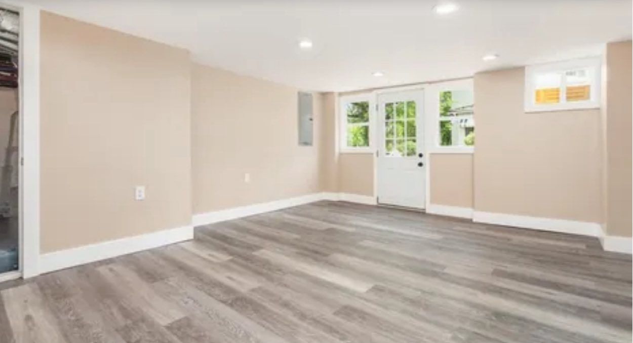 An unfurnished basement room with beige walls, grey wood-look flooring, recessed lighting, and a white exterior door.