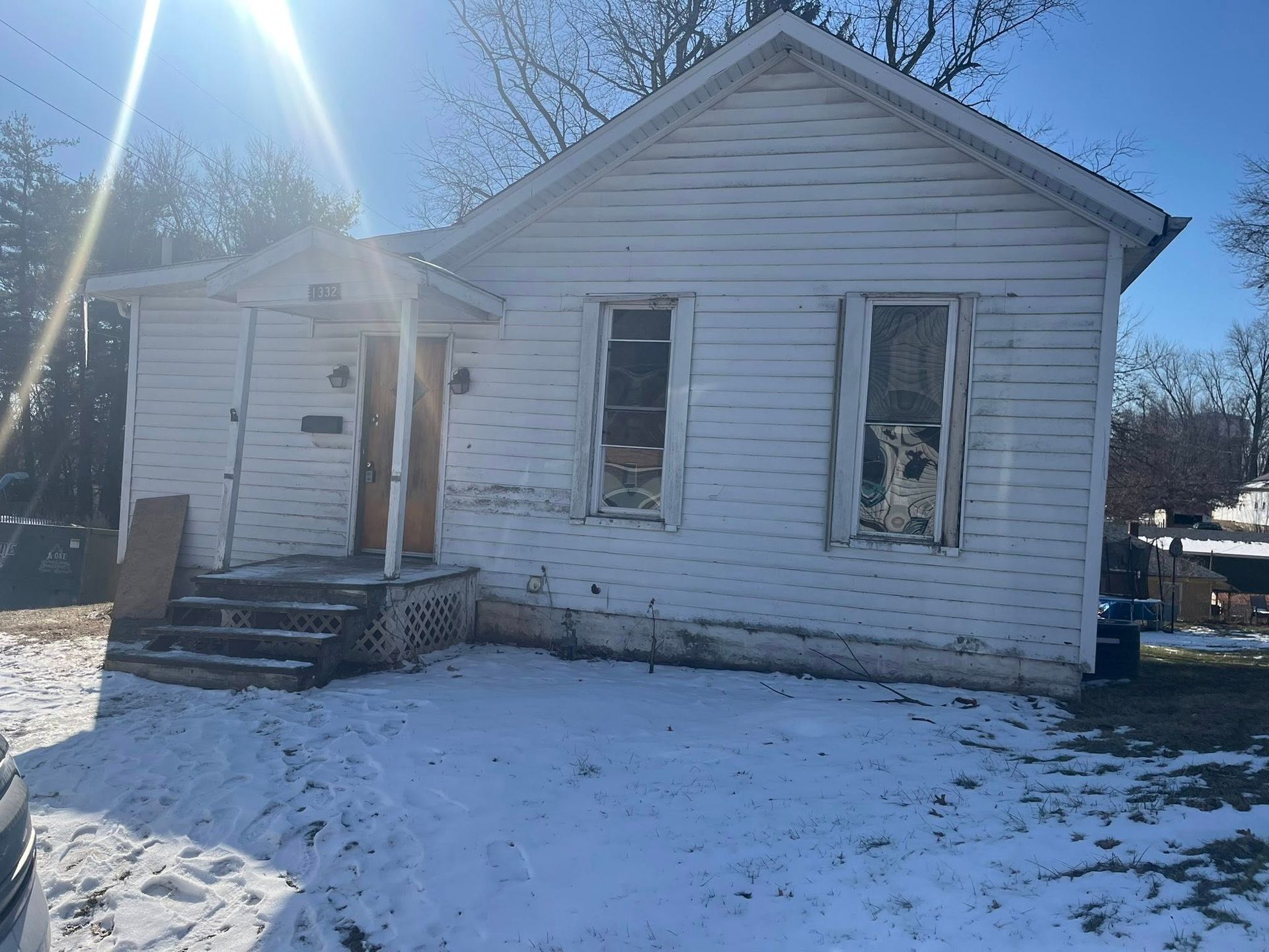 A small, weathered white wooden house with peeling paint, two front windows, and a small porch, set against snowy ground.
