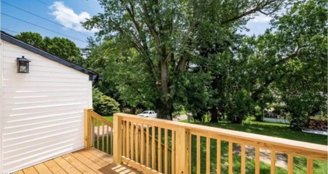 A view from a wooden deck overlooking a lush green yard with large trees and a white wall on the left.