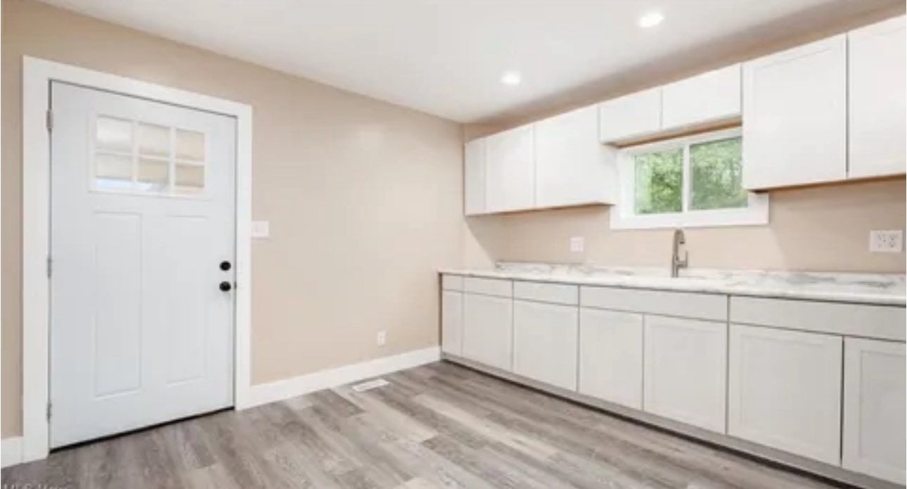 Modern kitchen featuring light-toned walls, grey wood-look flooring, white cabinets, and an exterior door.