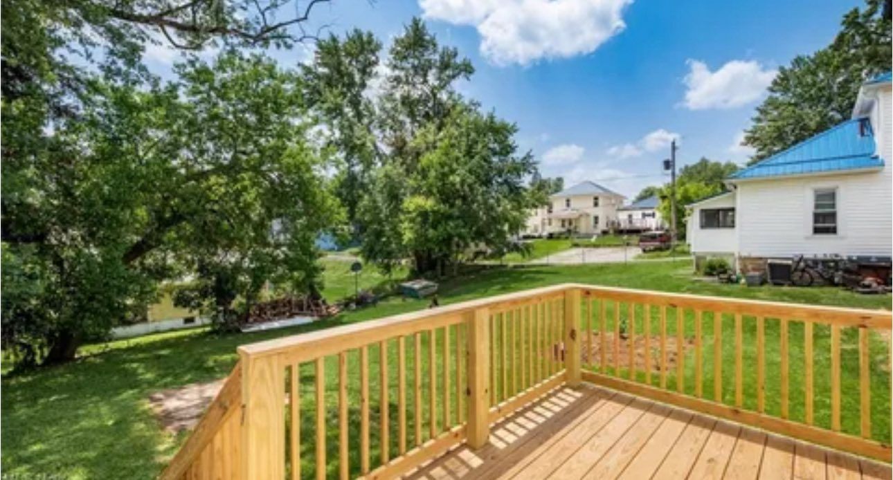 A wooden deck with railings overlooks a grassy backyard with trees and a white house under a blue sky.