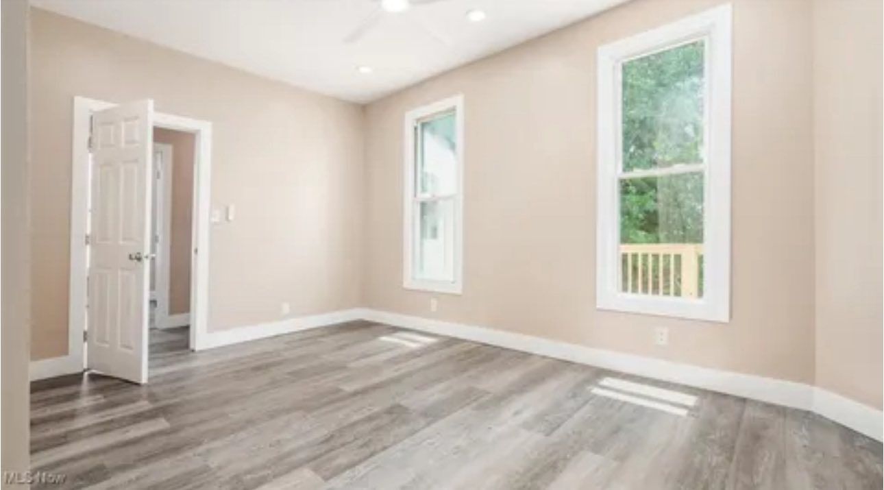 An empty bedroom with light beige walls, gray wood-look flooring, two white-trimmed windows, and an open white door.
