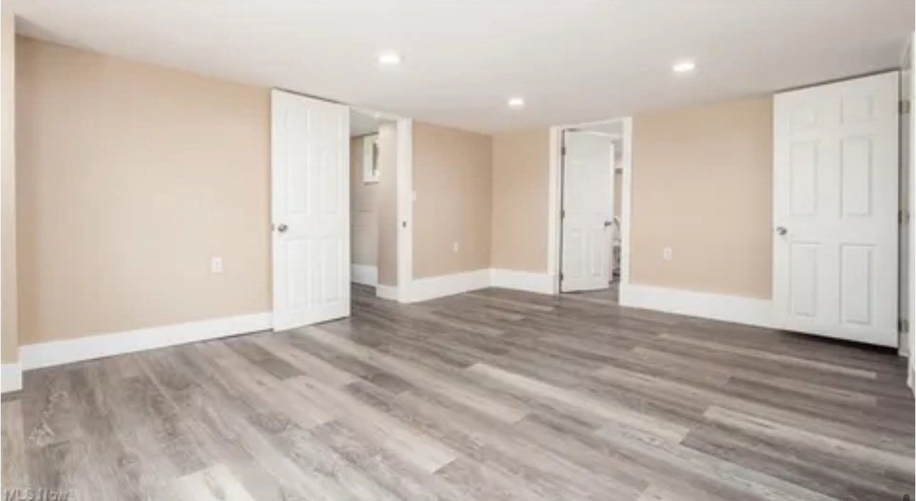 An empty room with beige walls, white trim, wood-look flooring, and three white doors leading to other areas.