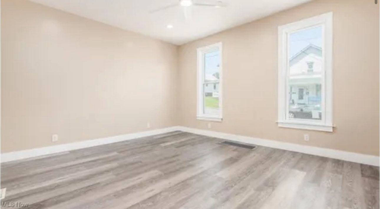 An empty room with light beige walls, gray wood-look flooring, two windows, and a ceiling fan.