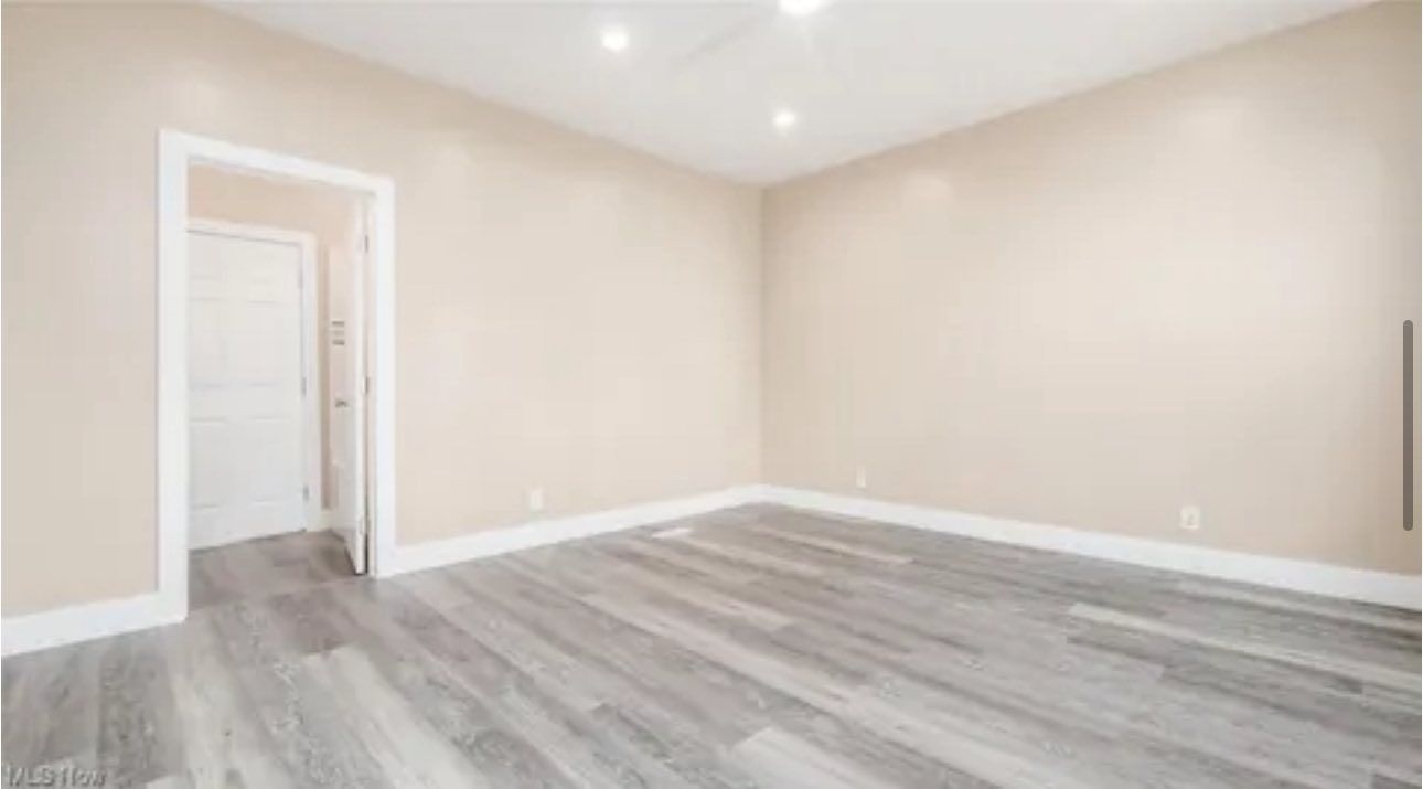 An empty, minimalist room with beige walls, grey wood-look flooring, recessed ceiling lights, and an open doorway.