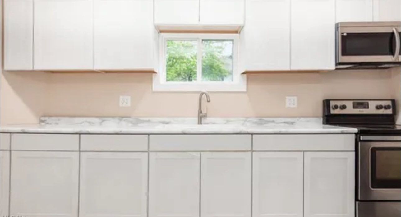 A kitchen features white cabinets, white marble-style countertops, a stainless steel stove, and a window above the sink.