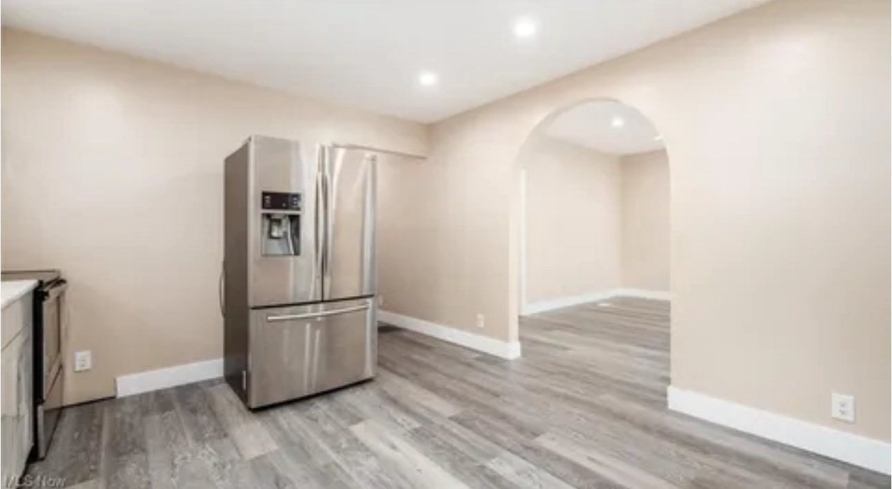 Stainless steel refrigerator in a room with light beige walls and grey wood-look flooring, next to an arched entryway.