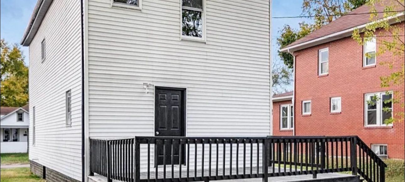 White house with a black door and porch railing, standing next to a red brick house under a blue sky.