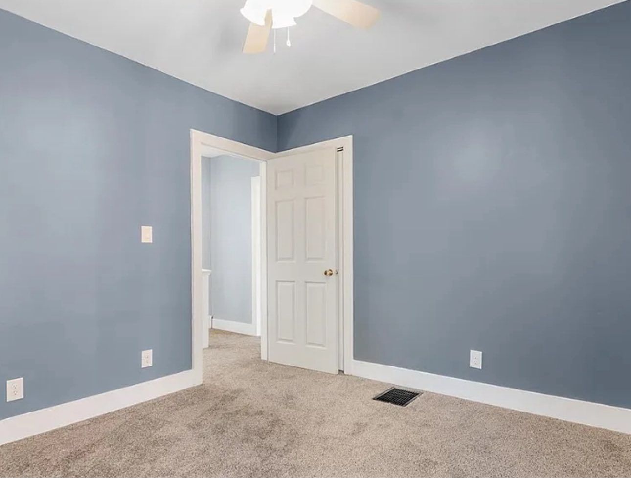 An empty room with light blue walls, beige carpet, white trim, a ceiling fan, and an open doorway leading to a hallway.
