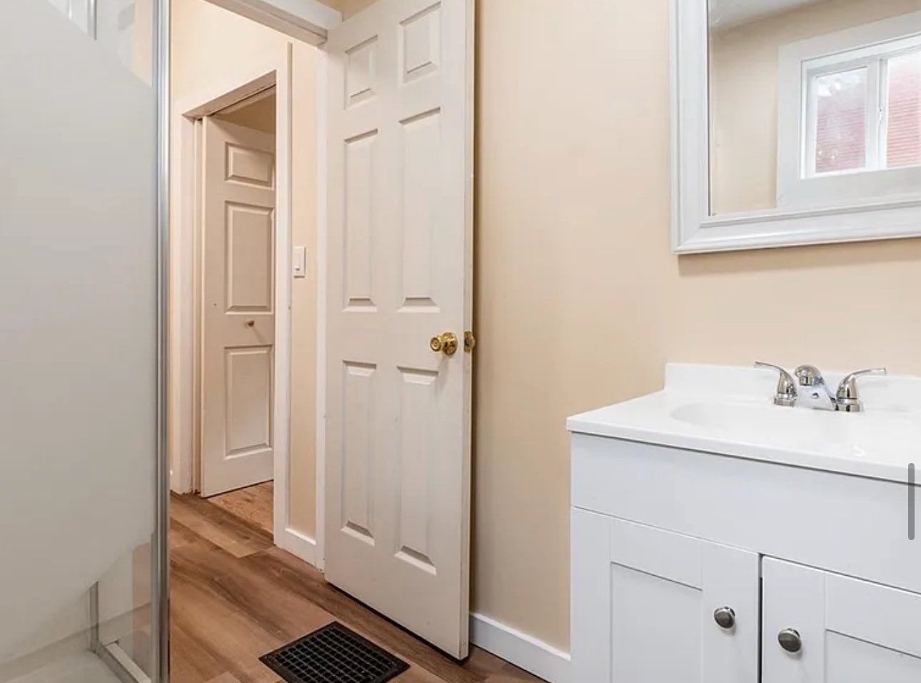 A modern bathroom featuring a white vanity with a chrome faucet, a wall mirror, and a beige door leading to a hallway.