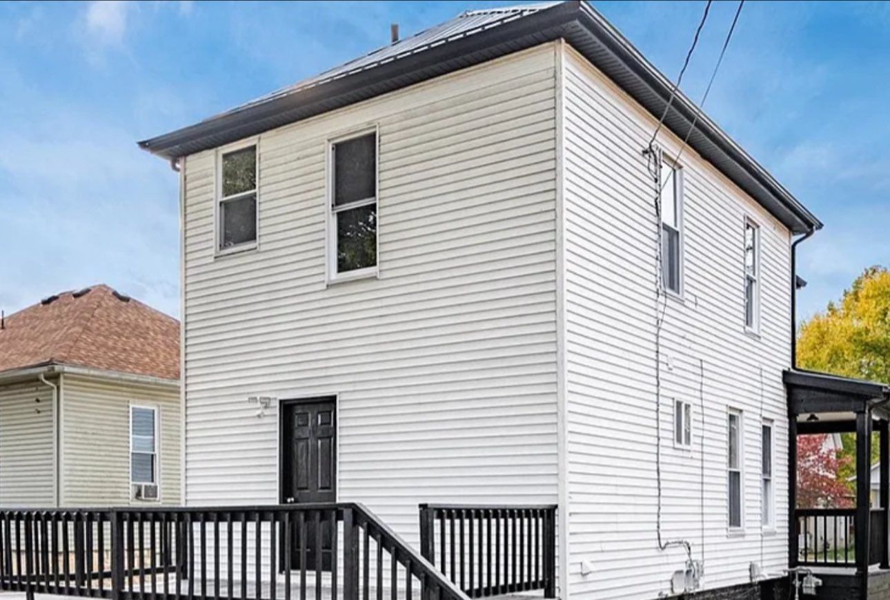 A two-story white house with light siding, dark trim, and a black deck against a blue sky.