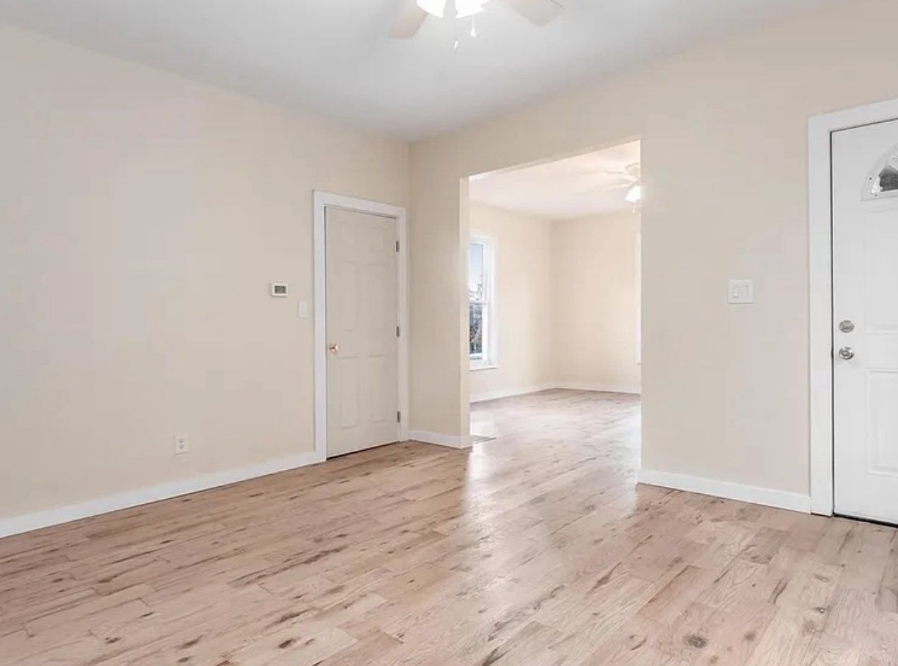 A bright, empty room with light-colored hardwood floors, beige walls, a single interior door, and an exterior front door.
