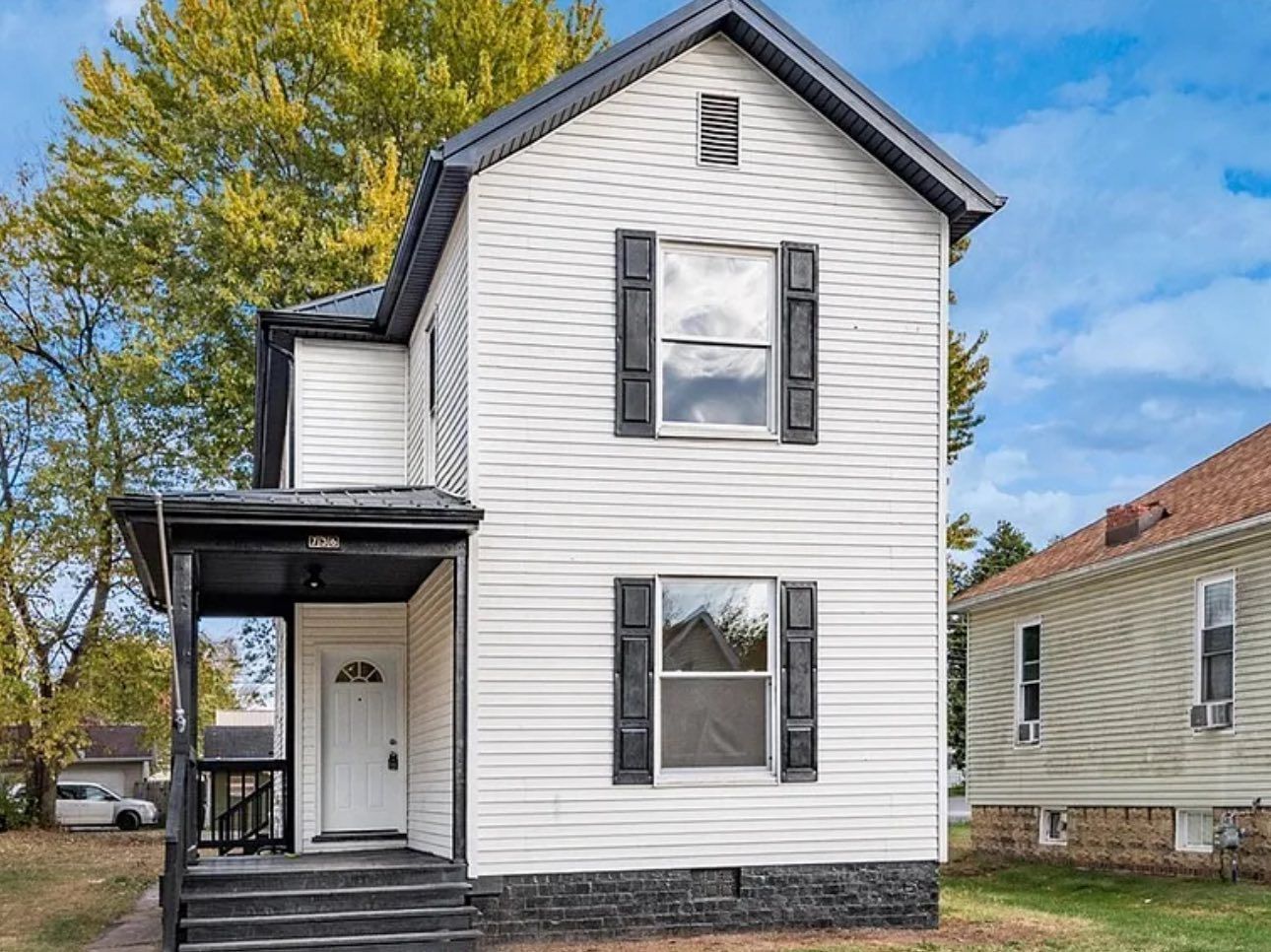 A two-story, white vinyl-sided house with a black front porch, dark window shutters, and a stone foundation.