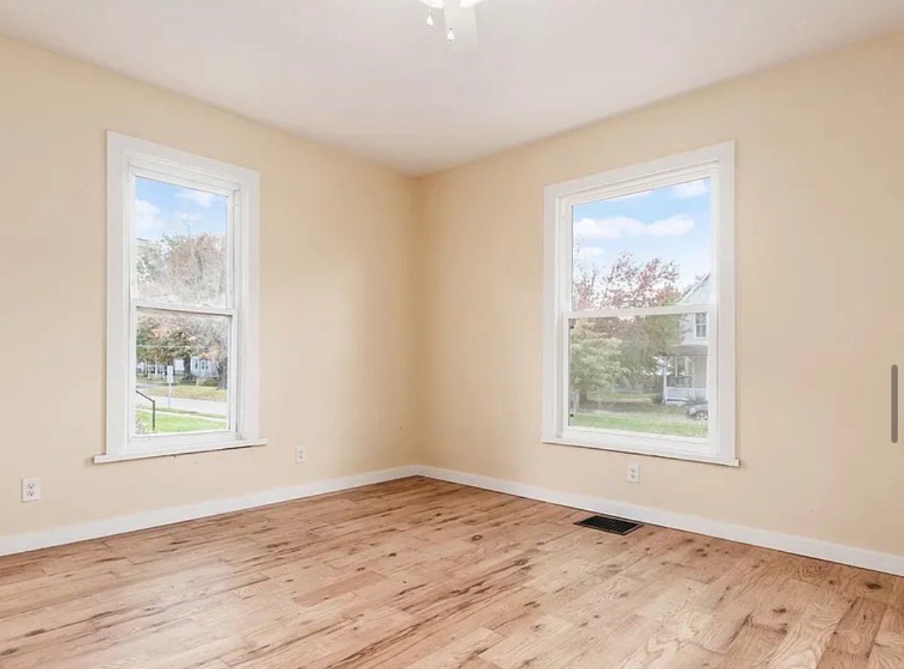 An empty room with light beige walls, hardwood flooring, and two white-framed windows overlooking a suburban street.