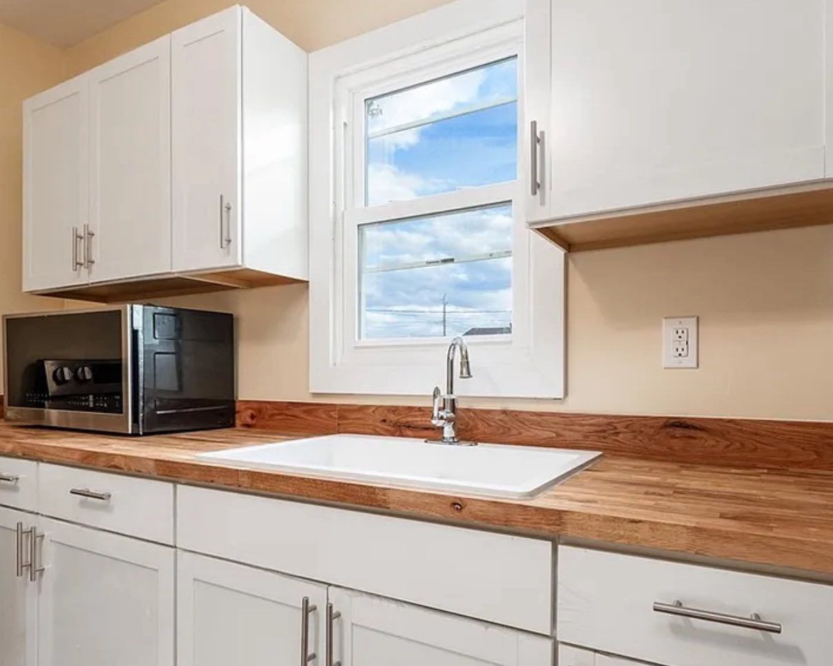 White kitchen cabinets with silver pulls, wood countertop, a white sink with a faucet, and a microwave by a window.