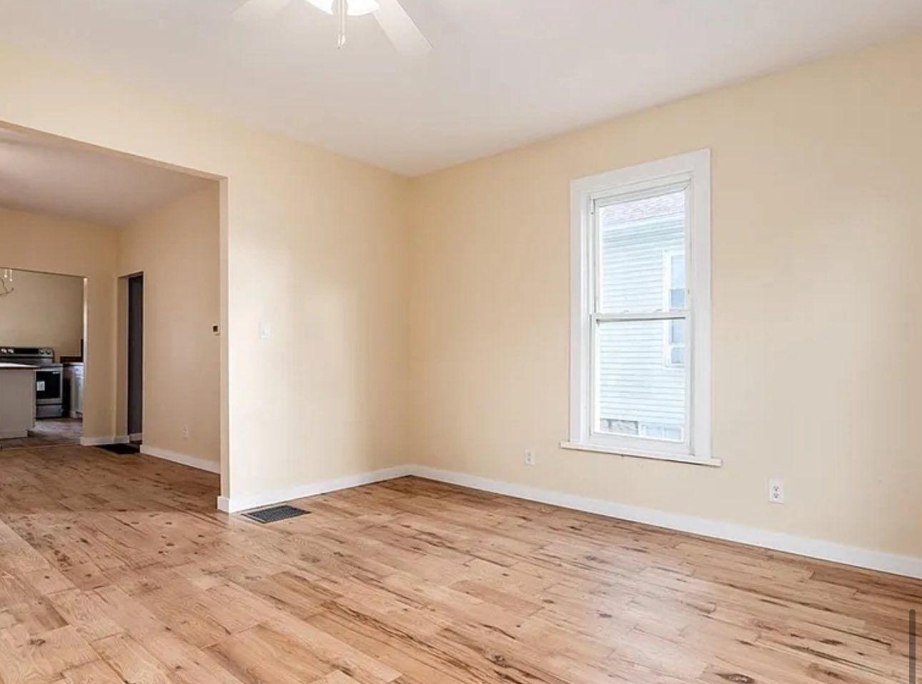 A bright room with light tan walls, hardwood-style flooring, a single window, and an open doorway to a kitchen.