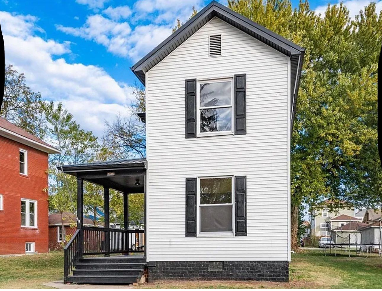 Two-story white house with black shutters and a front porch, located next to a red brick building on a sunny day.