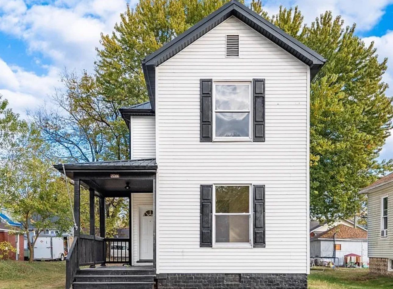 A two-story, white-sided house with a black roof, black window shutters, a front porch, and a dark foundation.