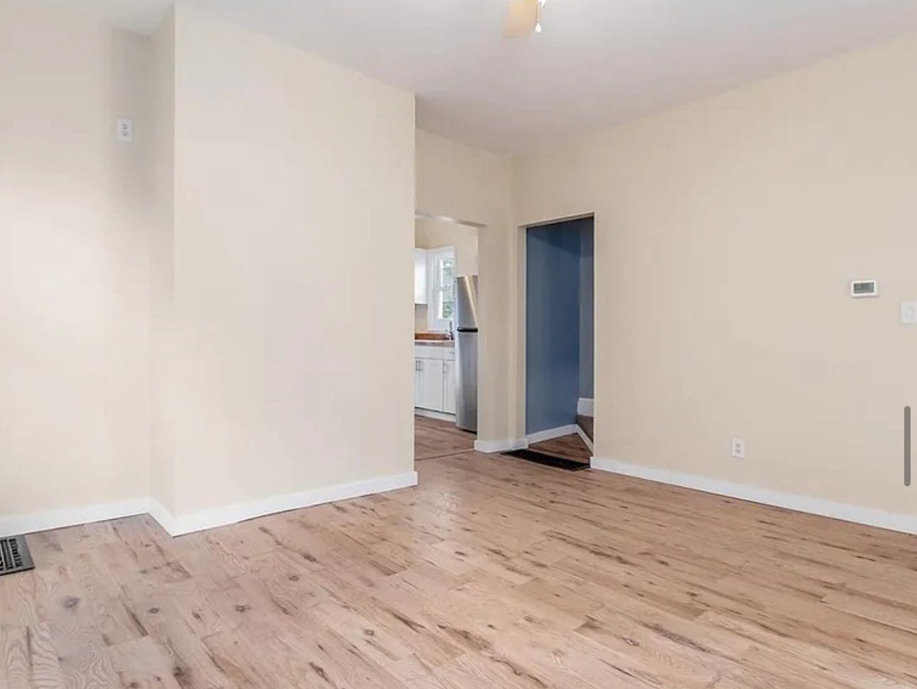 An empty room with light wood floors and cream-colored walls, featuring a doorway into a kitchen area.