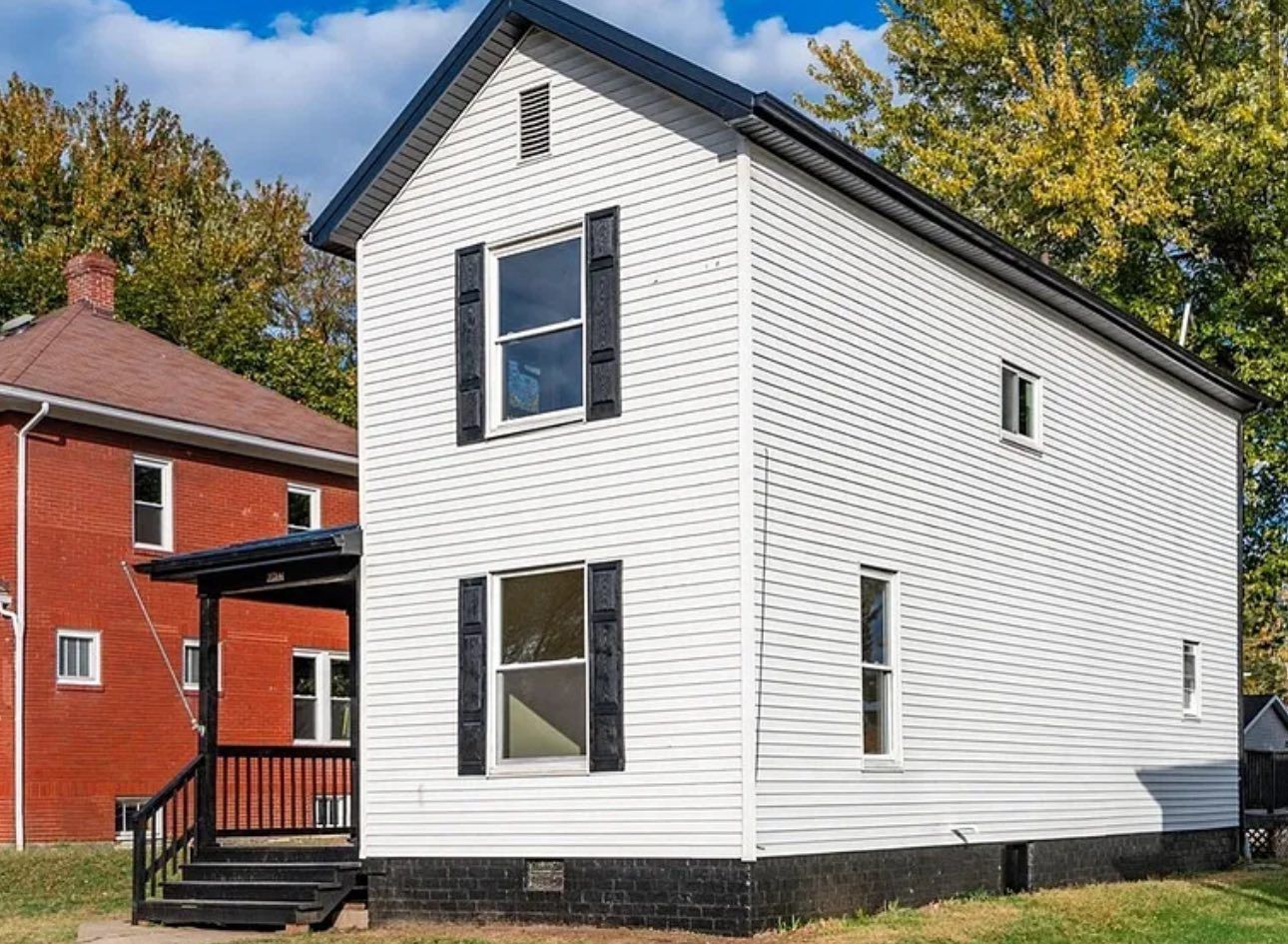A narrow, two-story white house with black shutters and a small front porch, situated next to a red brick home.