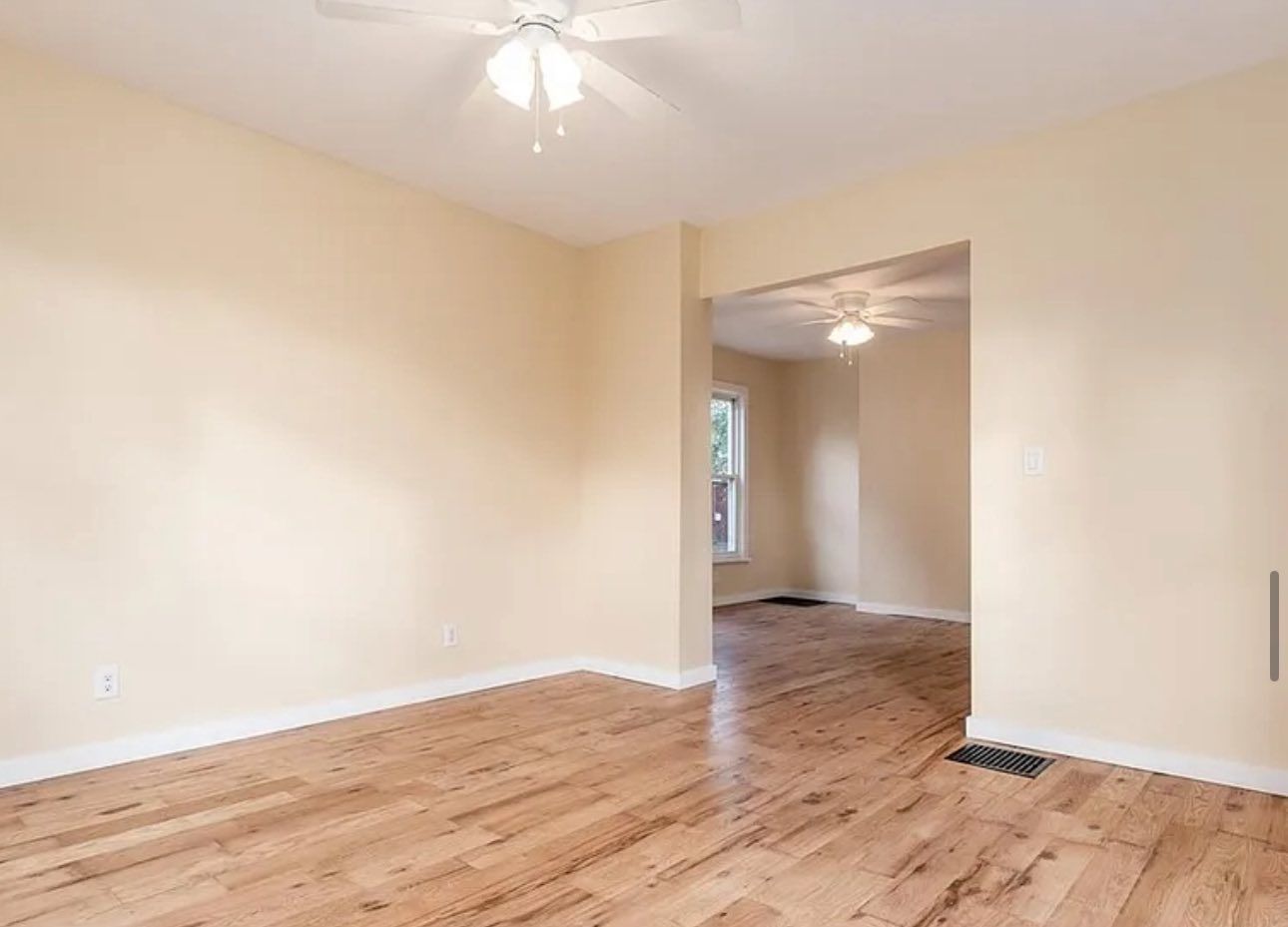 An empty, light-walled room with hardwood flooring, a ceiling fan, and a wide doorway leading to an adjacent area.