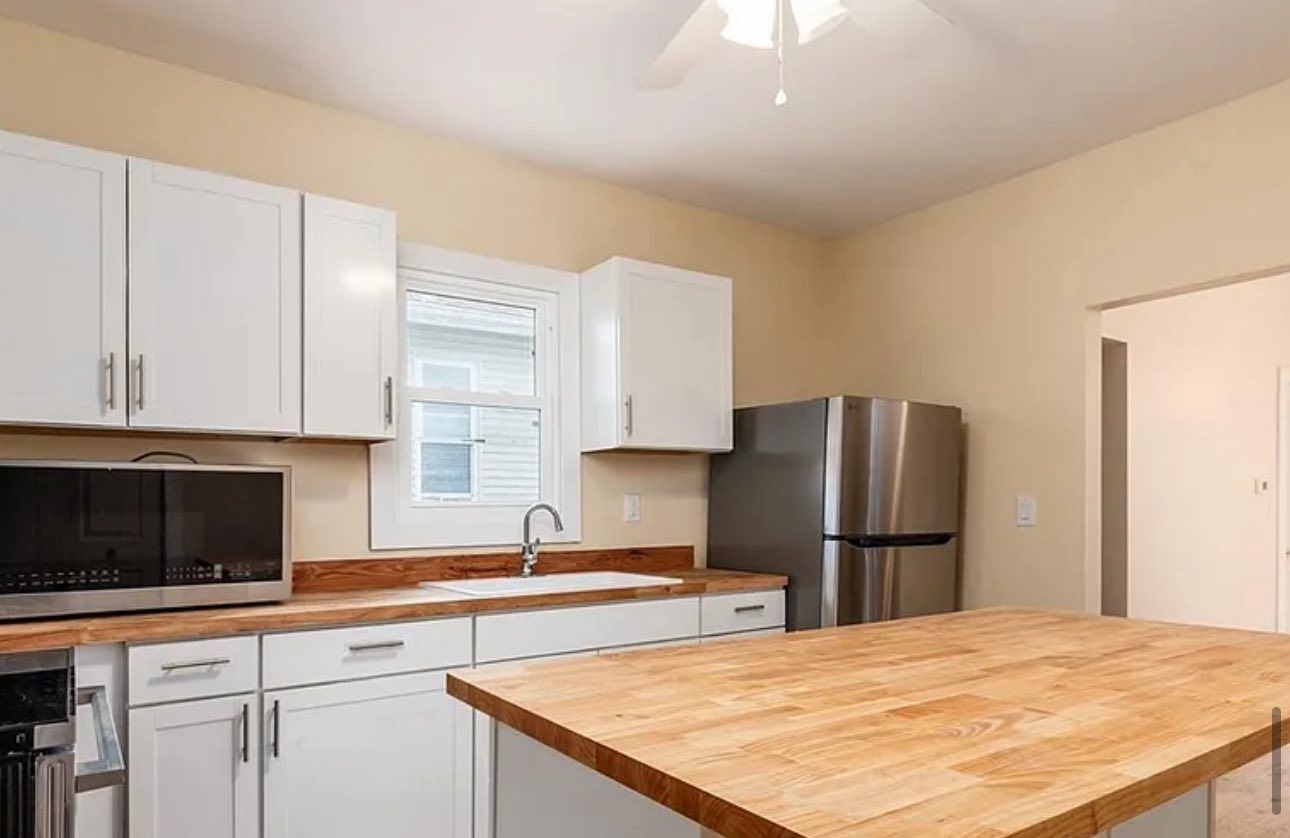 A modern kitchen featuring white cabinets, wood countertops, a stainless steel microwave and refrigerator, and a window.