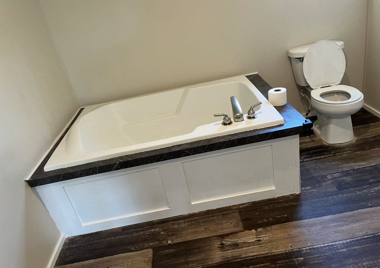A white drop-in bathtub with black trim and a white toilet in a bathroom with dark wood-look plank flooring.