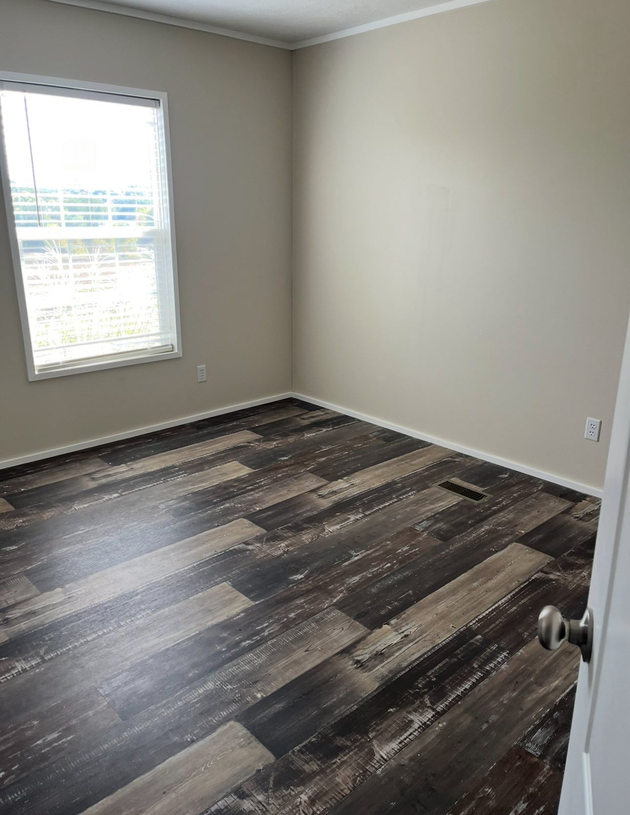An empty room with light beige walls, a window on the left, and dark, wood-look plank flooring.