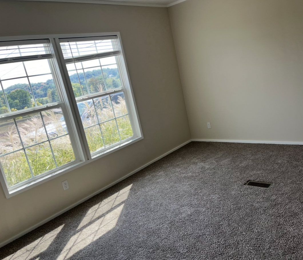 An empty, beige room with gray carpet, a double-window overlooking a construction site, and a floor vent.