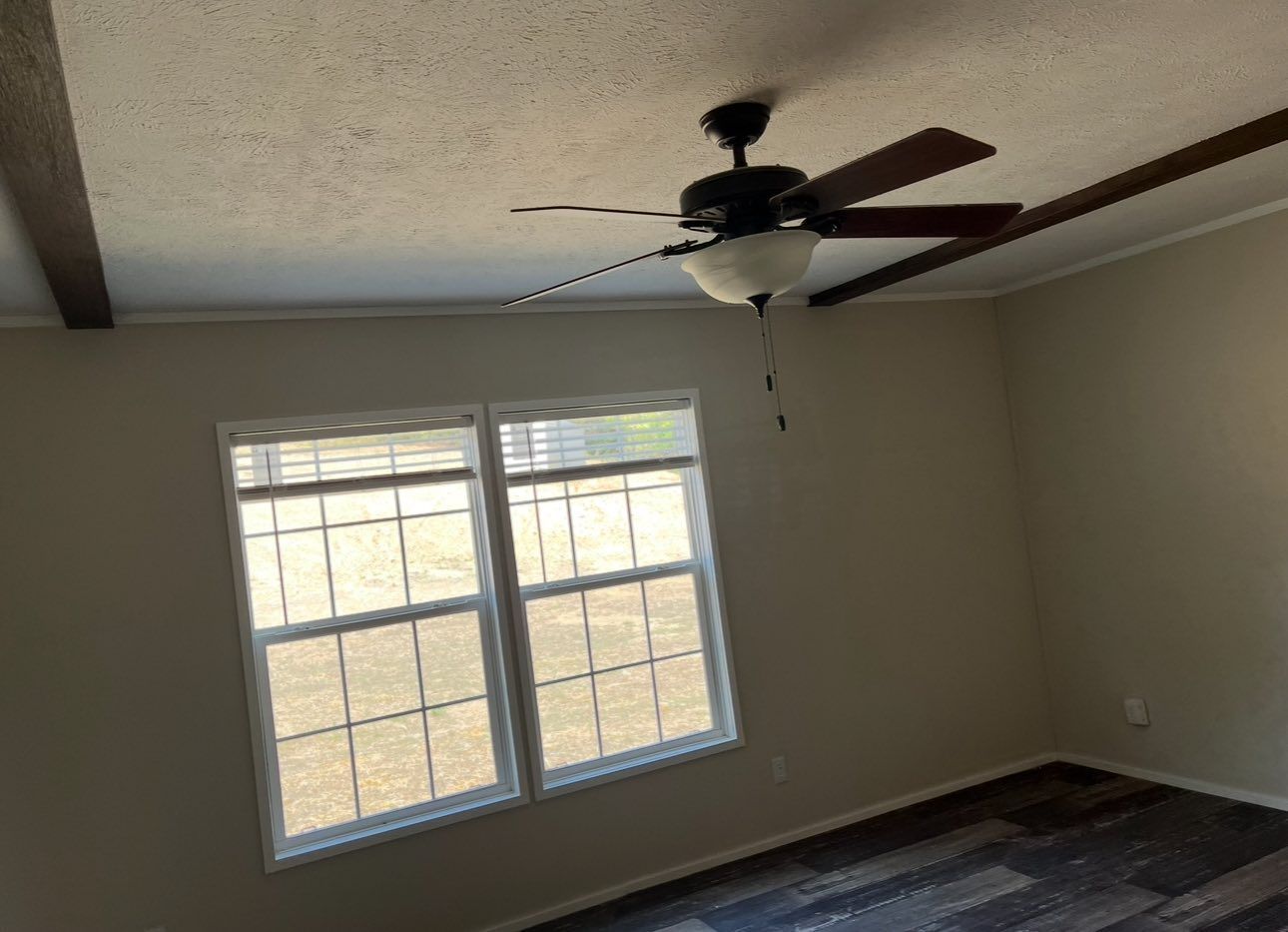 An interior room with beige walls, a popcorn ceiling with wood-look beams, a ceiling fan, and a window with white blinds.