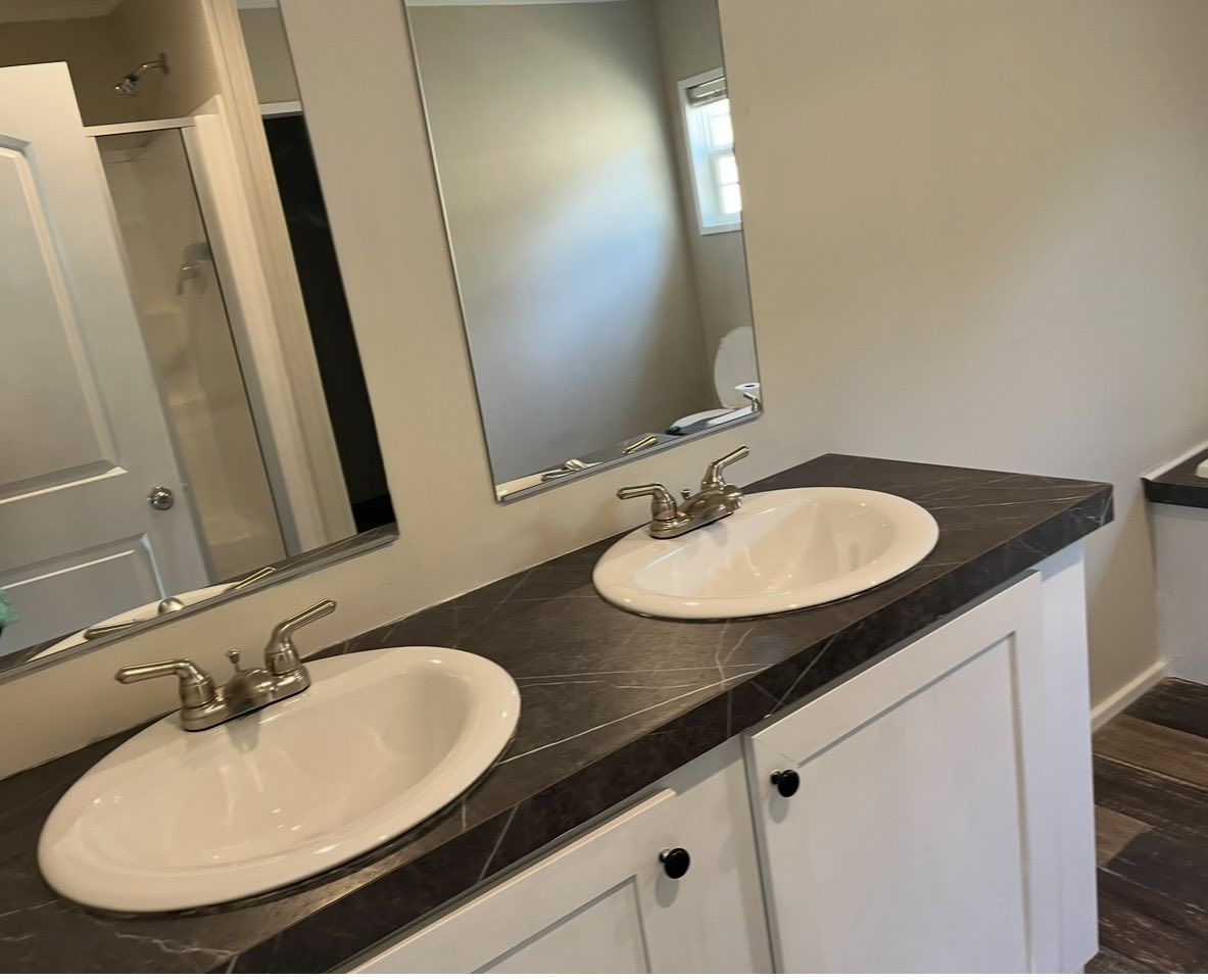 A bathroom vanity with two white oval sinks set into a dark, faux-marble countertop with white cabinets and wall mirrors.