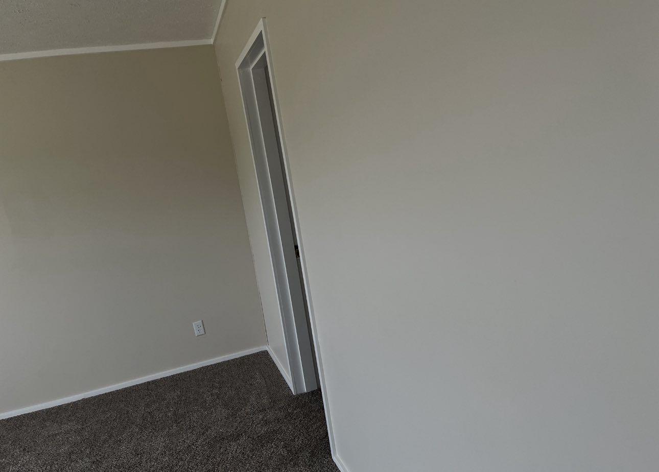 An interior corner of a room with light beige walls, brown carpet, and an open doorway leading to a hallway.