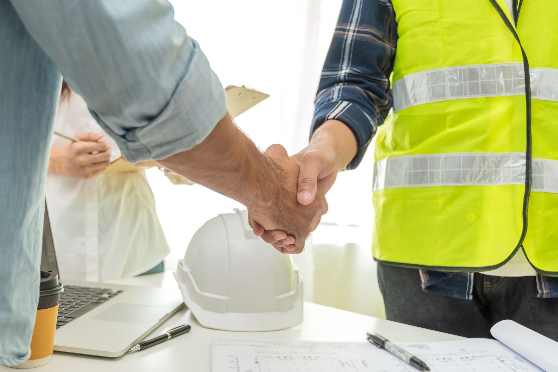 A handshake between a contractor and a client over a desk with a hard hat, plans, and laptop nearby