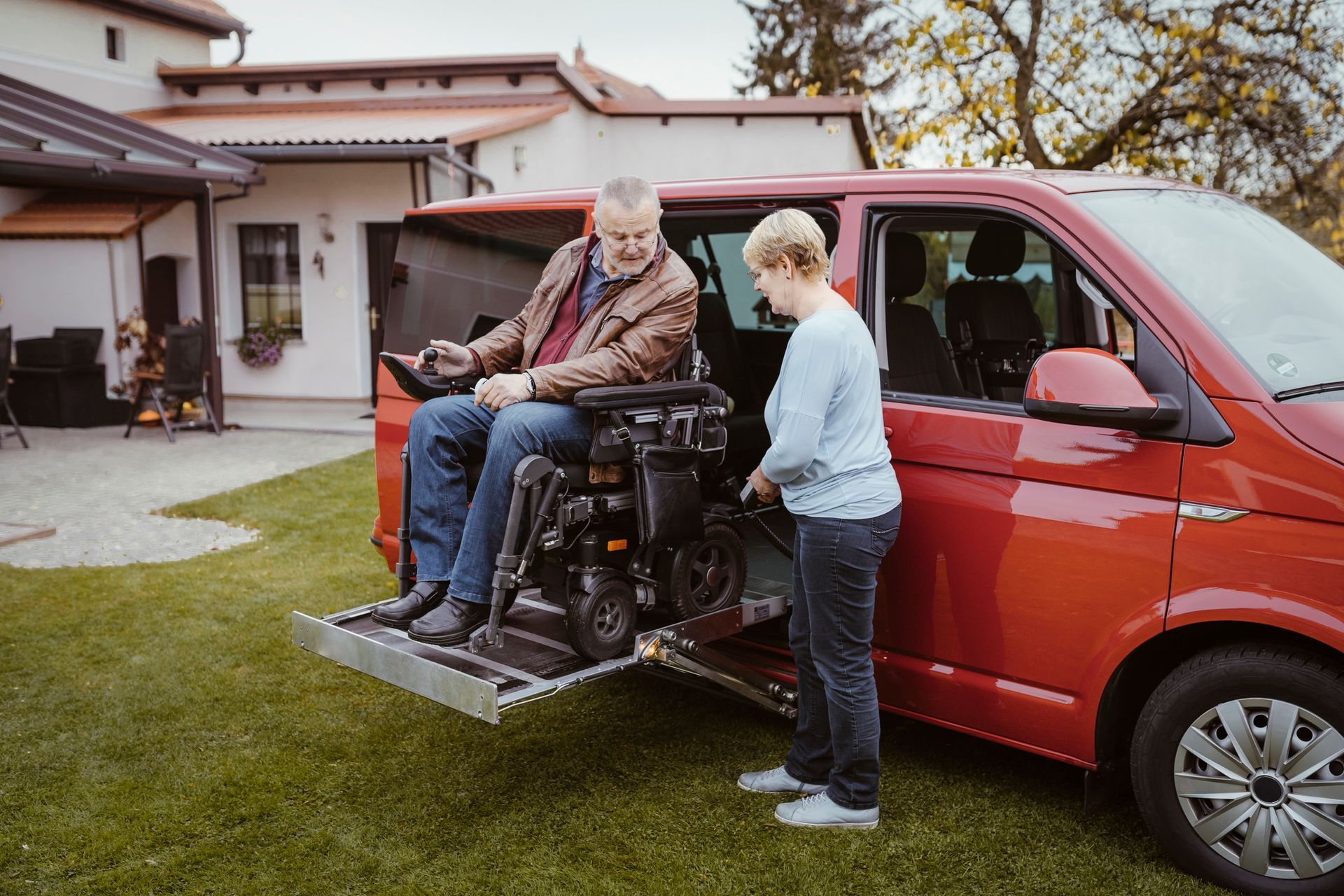 Elderly man in a wheelchair getting into a vehicle with a motorized lift by a woman.