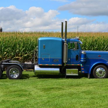 A blue semi truck is parked in a grassy field with corn stalks behind it