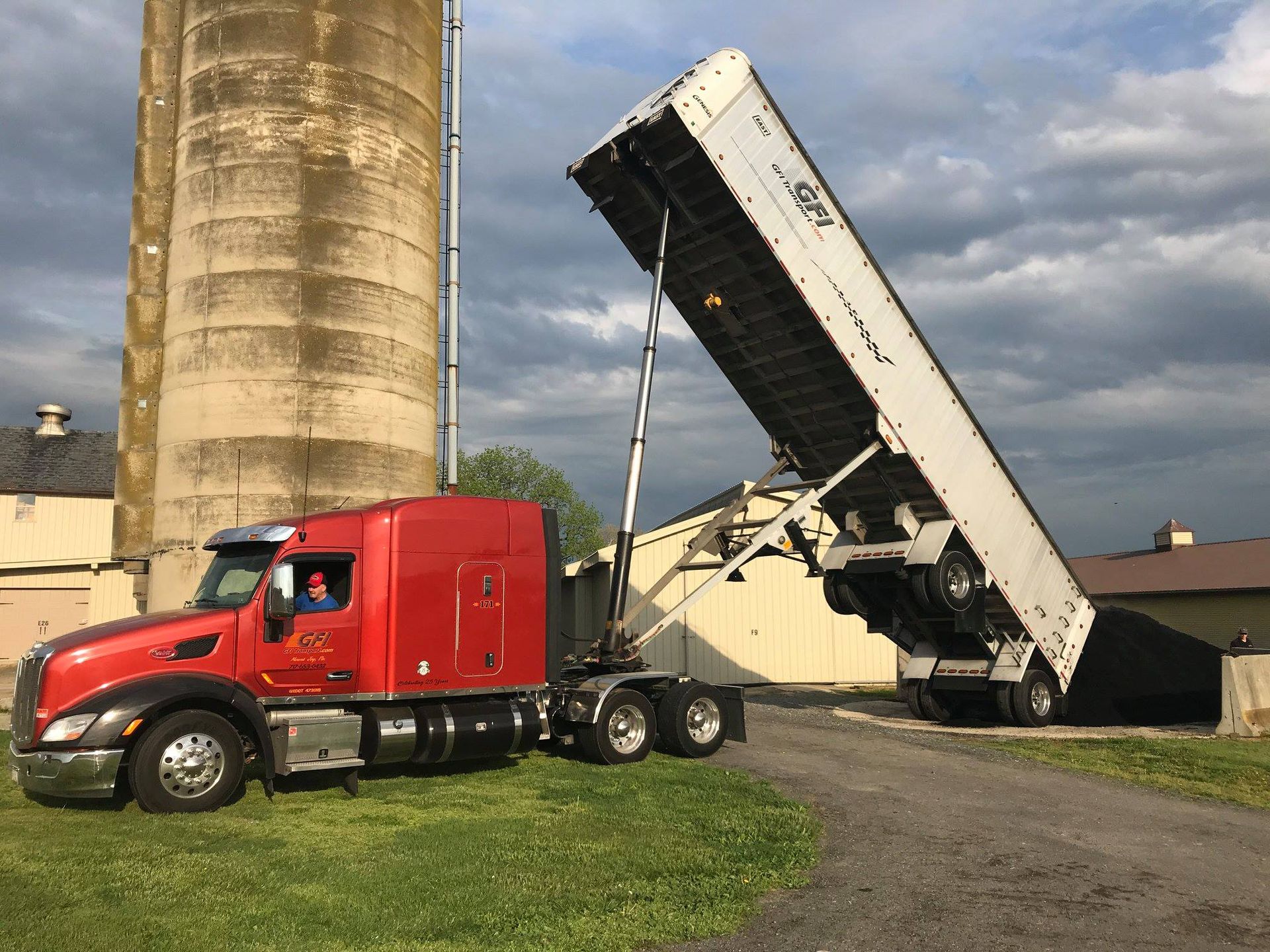 Red semi-truck tilting trailer up, unloading contents near a silo. Driver in cab. Green grass and light sky.