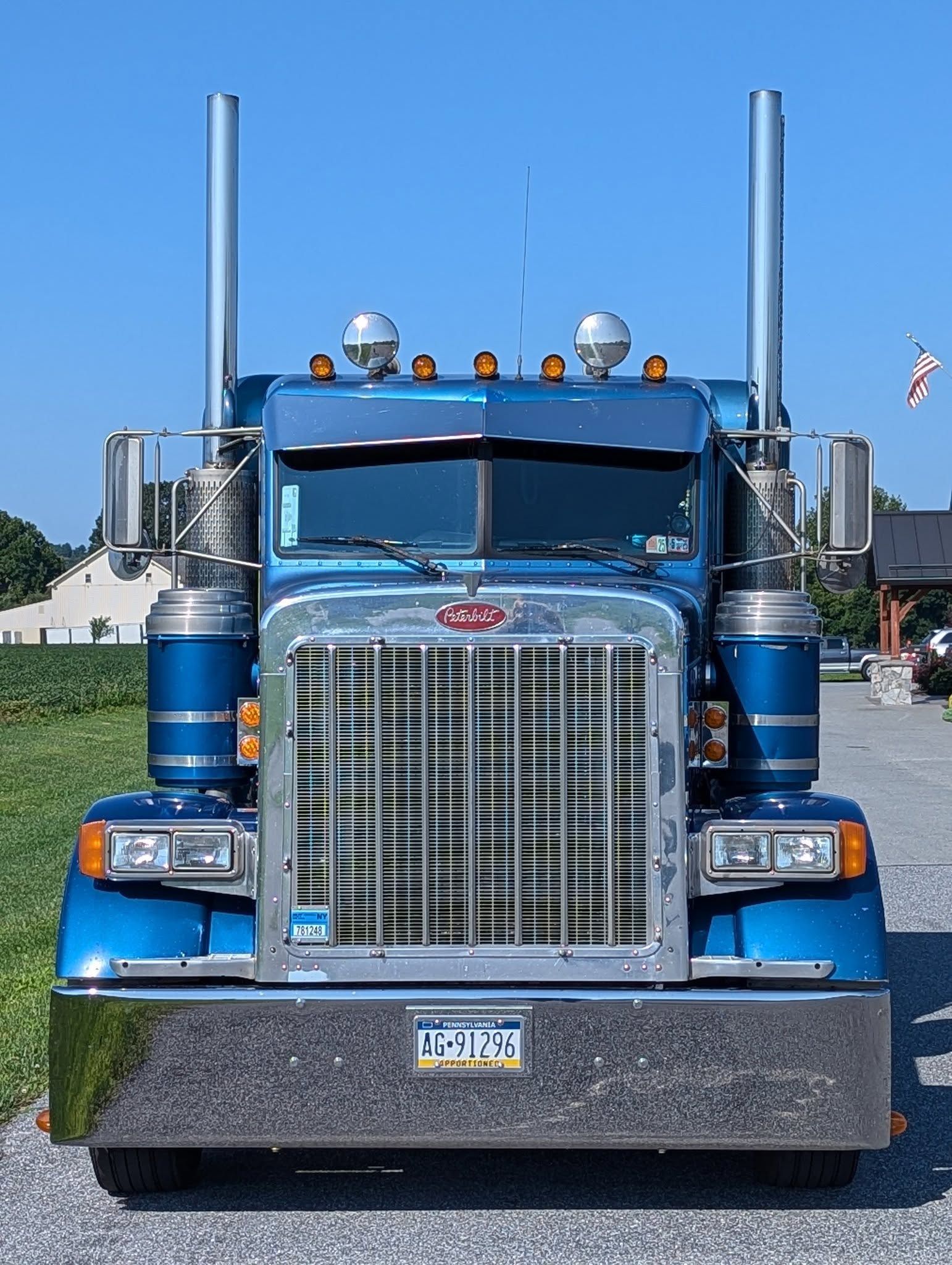 A blue Peterbilt semi-truck viewed from the front, featuring a large chrome grille and two prominent vertical exhausts.