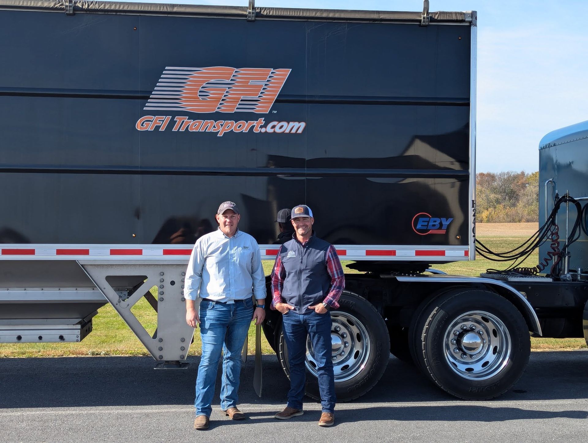 Two men stand in front of a black trailer truck with GFI Transport logo. Sunny outdoor setting.