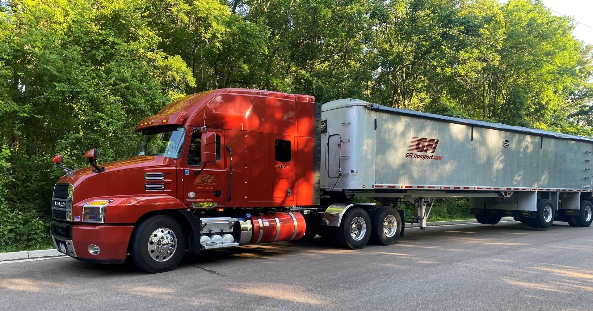 A bright red Mack semi-truck with a silver trailer parked on a road with green trees in the background.