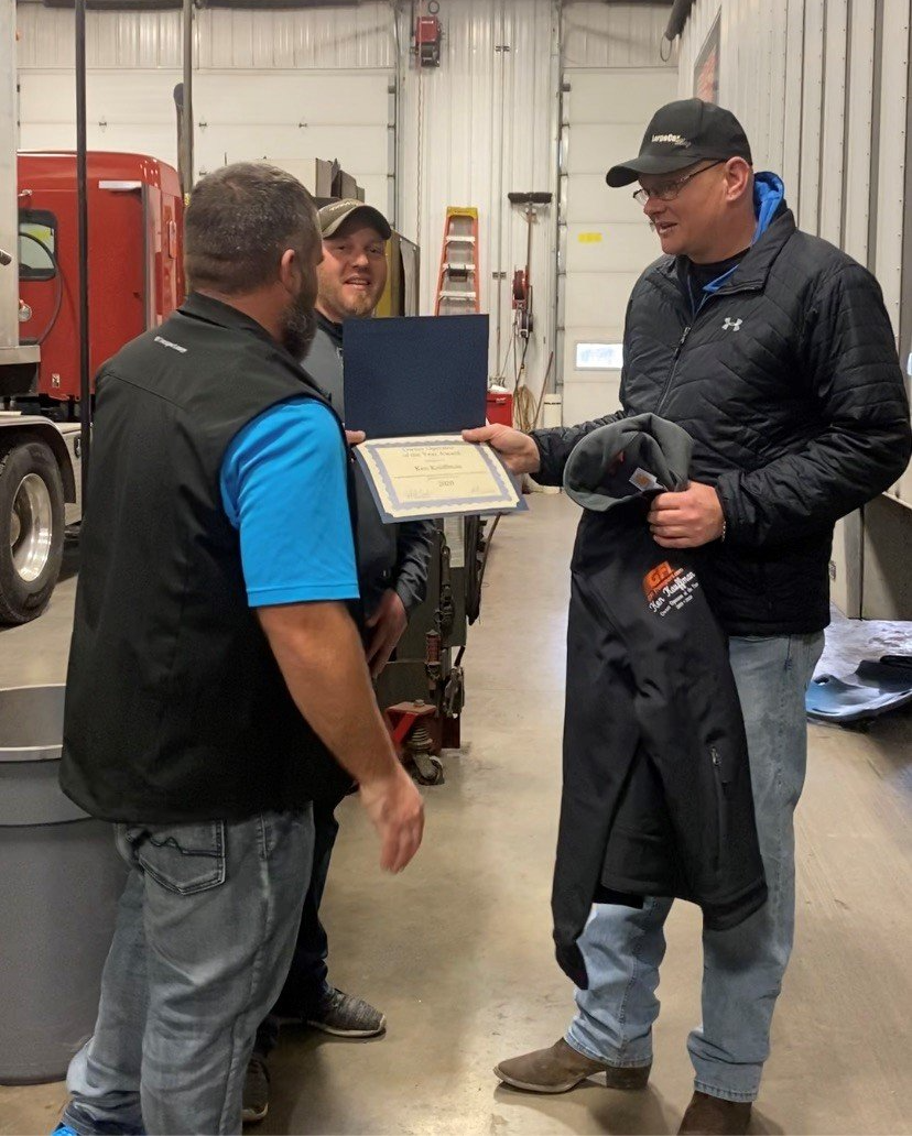 Three men are standing in a garage and one of them is holding a certificate