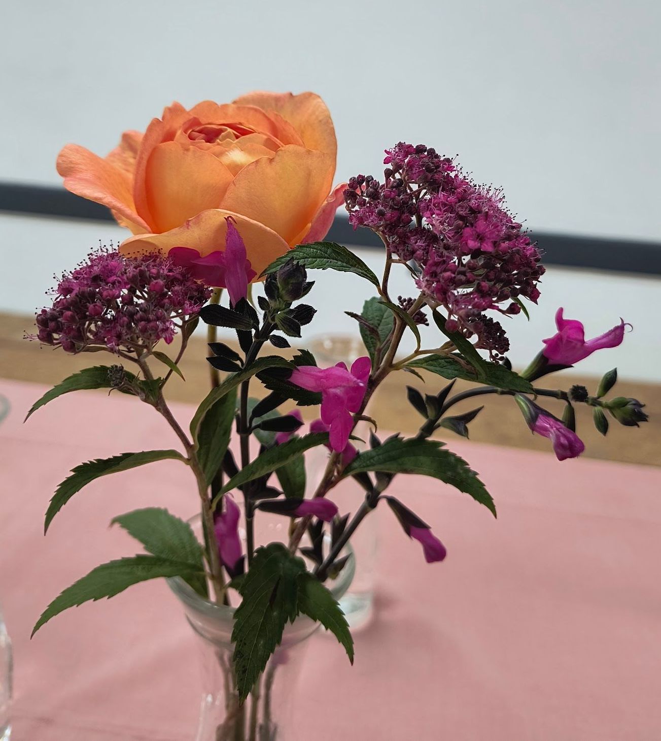 Image shows a floral arrangement in a glass vase comprising a peach coloured rose, 2 sprigs of dark pink Spirea japonica and 2 sprigs of dark pink salvia