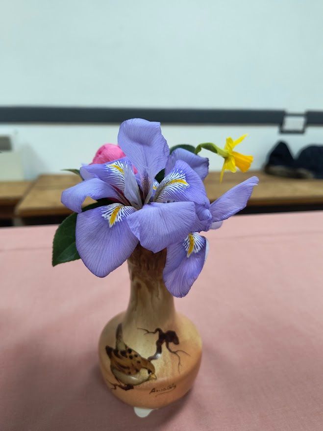 Photo shows blue irises and one daffodil in wooden vase with bird decoration