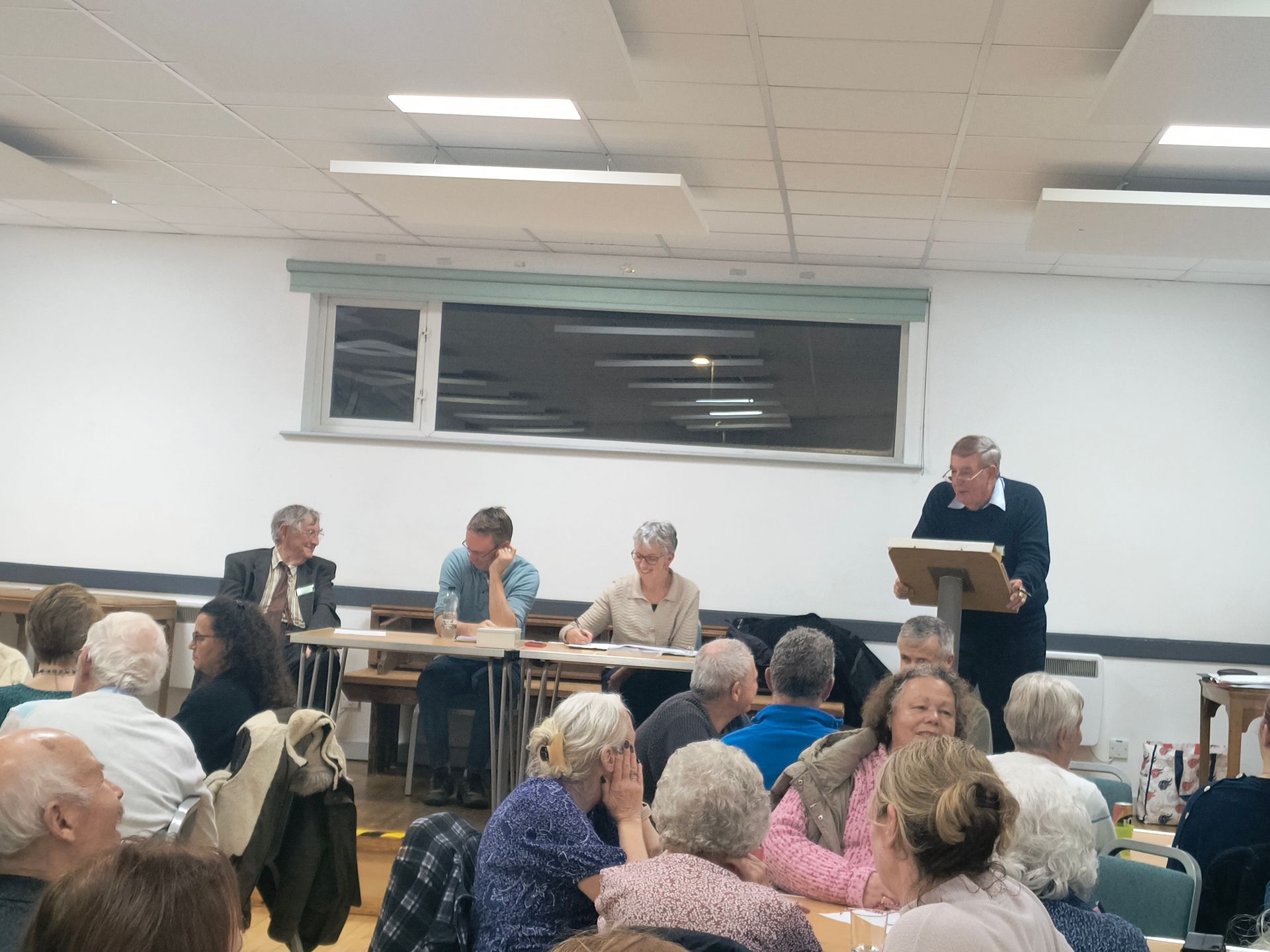 Club Chair Glenn Duggan stands at a podium, 3 members of the committee sit next to him on the stage, some members seated at tables off the stage are visible in the foreground
