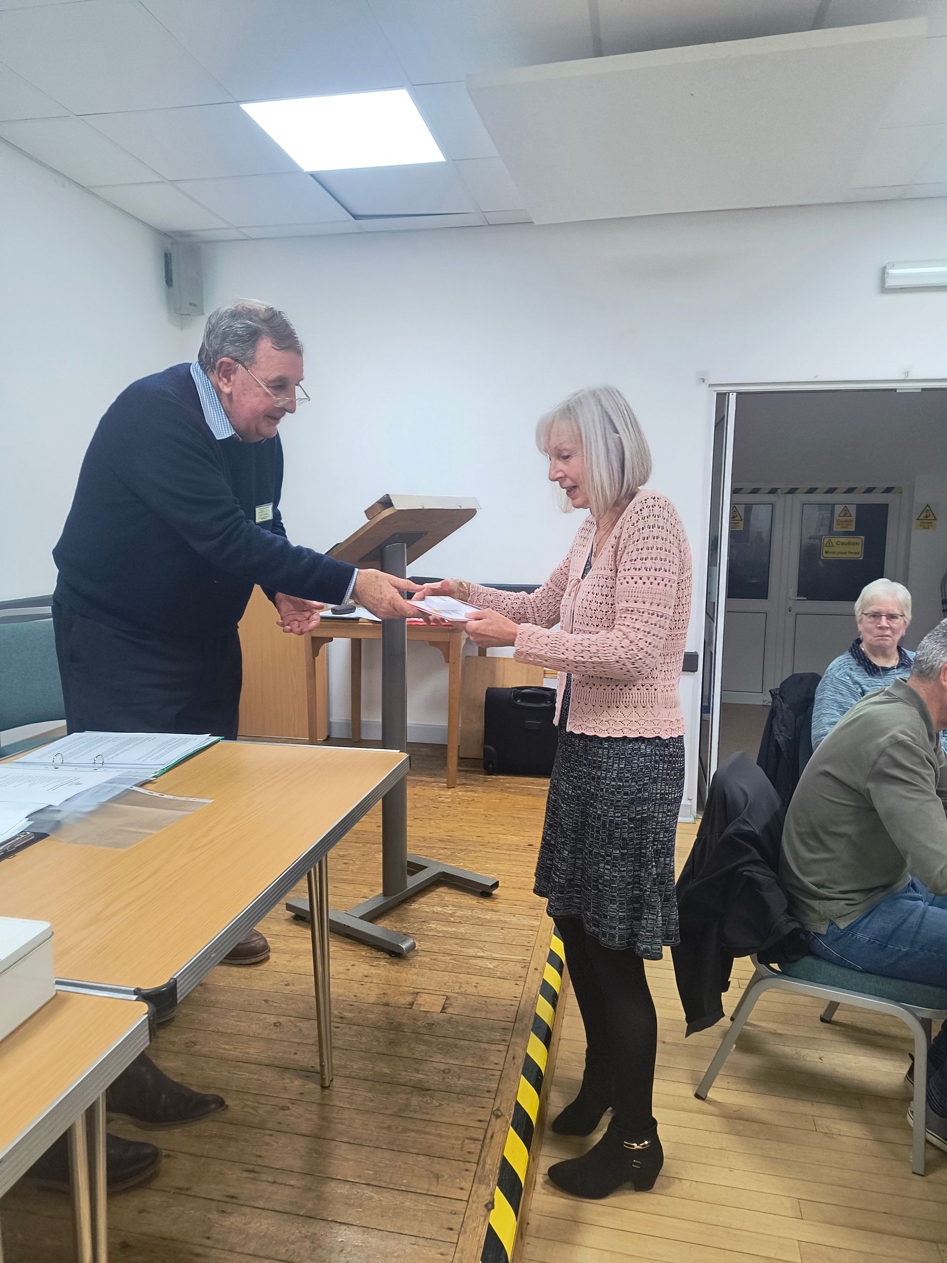 Glenn Duggan stands on the stage passing a box and certificate to Lynne Pederson who is in front of the stage
