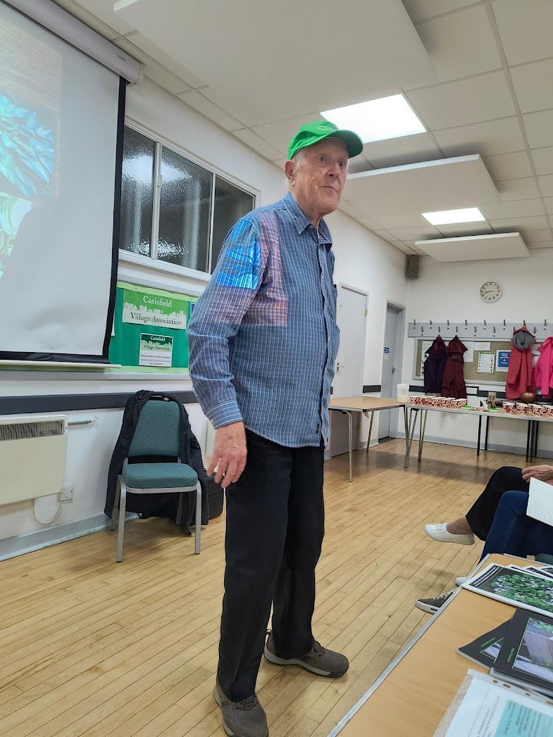 Photo shows John Barker wearing a striped blue shirt and green cap standing in front of the projection screen at the front of the room