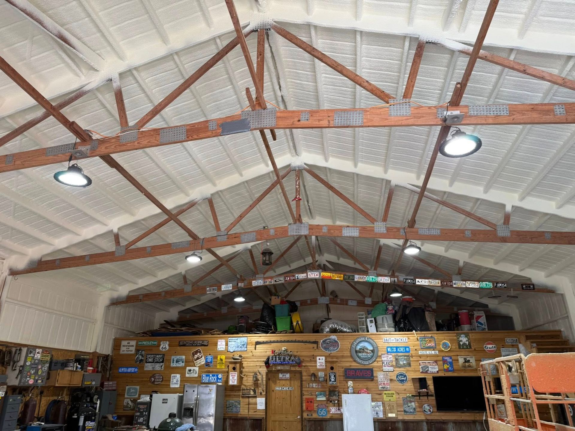 Interior view of a garage with spray foam insulation on ceiling and walls. Overhead lighting, wooden beams, and a wall of items.