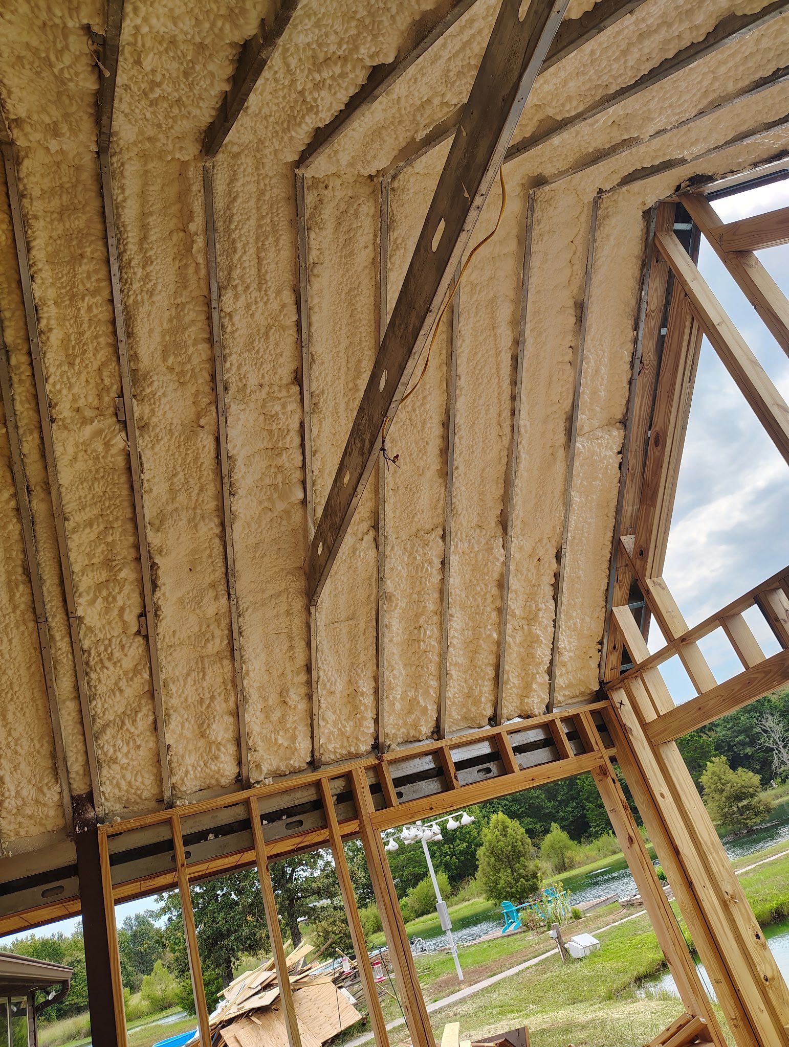 Spray foam insulation applied to the underside of a roof's wooden structure, outdoors with a partly cloudy sky.