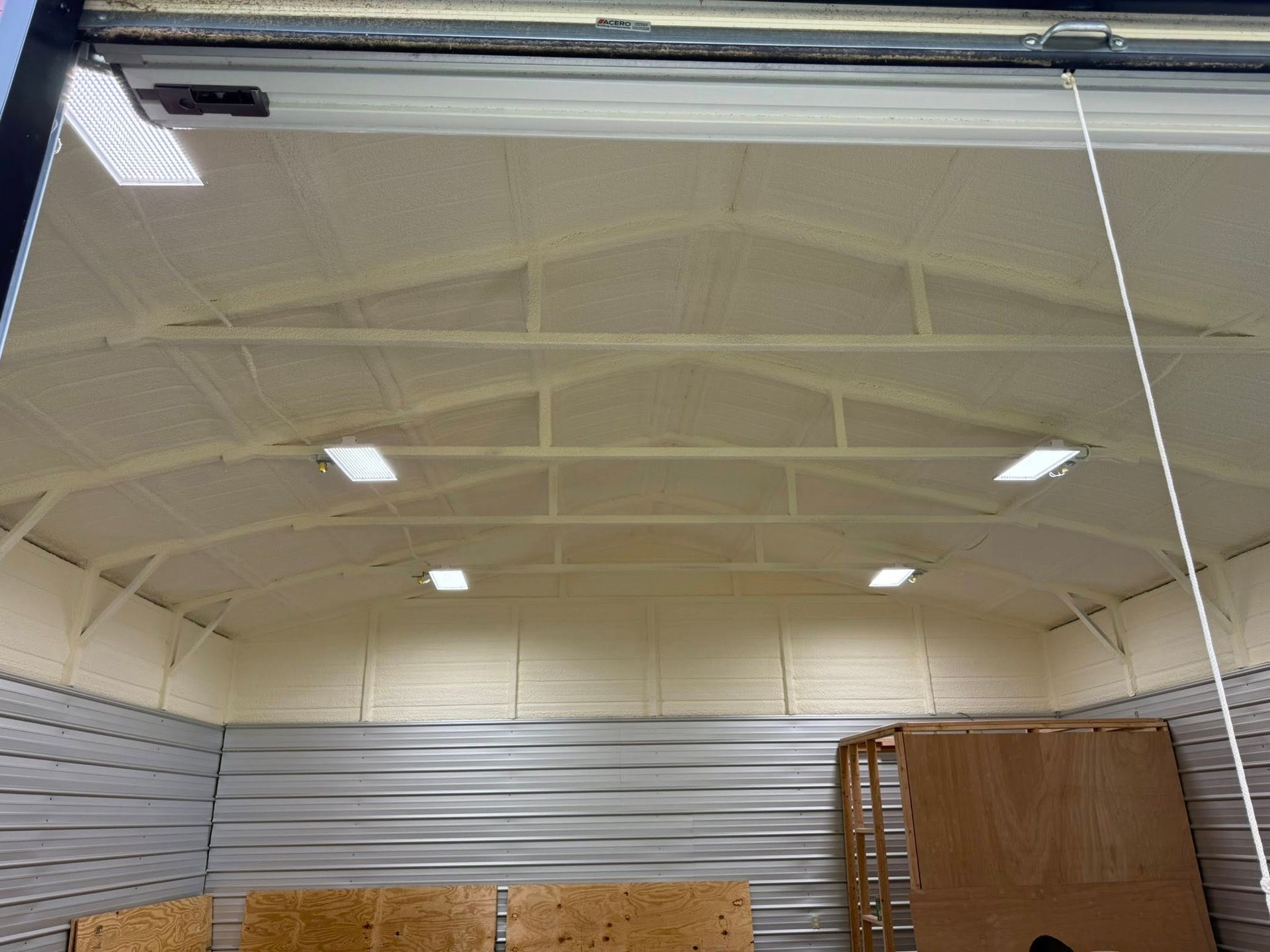 Interior view of a metal building with spray foam insulation, overhead lights, and a partially open door.